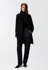 Cappotto di lana nero con colletto rotondo e chiusura con bottoni, indossato sopra una blusa nera trasparente e pantaloni neri su misura, abbinato a tacchi a punta neri.