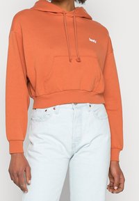 Cropped orange hoodie with a front pocket, drawstrings, and white logo. Paired with light blue high-waisted jeans. Soft texture.
