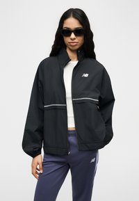 Γυναίκα που φοράει μαύρο μπουφάν με φερμουάρ, λευκό crop top, μοβ παντελόνι New Balance και μαύρα γυαλιά ηλίου, στέκεται μπροστά σε απλό φόντο.