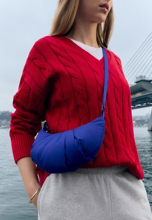 Woman wearing red cable knit sweater and gray pants, carrying a blue padded crossbody bag, standing near water with a bridge in background.
