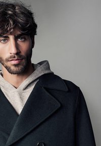 Schwarzer Wollmantel mit Reverskragen, doppelt knöpfbarem Verschluss, darüber getragen ein hellgrauer Kapuzenpullover mit Kordelzug.