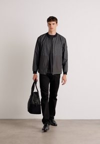 Black bomber jacket with a textured upper, paired with a black T-shirt, black pants, and carrying a black bag, walking on a light floor.
