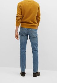 Yellow textured sweater, blue jeans, slim fit with back pockets, worn with dark shoes. Plain background.