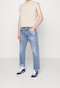 Beige ärmlös topp, ljusblå jeans med rak passform och fransad kant, kombinerat med svarta och vita sneakers.