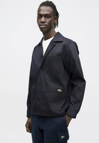 Chaqueta de algodón azul marino con cuello, cierre de botones y dos bolsillos delanteros. Cuenta con un pequeño logo verde de Lacoste en el bolsillo izquierdo.