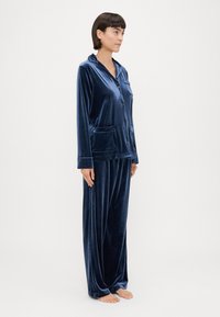 Conjunto de pijama em veludo azul-marinho, com blusa de botão, gola, mangas longas, bolso no peito e calças largas a condizer.