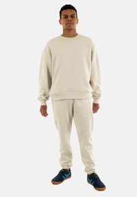 Felpa in cotone beige con maniche lunghe, polsini a costine e pantaloni della tuta svasati abbinati, accompagnati da scarpe scure con dettagli blu.