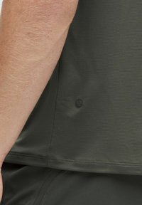 T-shirt de sport vert foncé en tissu texturé, avec un détail de logo sur le côté et un ourlet lisse. Texture des bras visible.