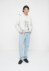 Haut à capuche gris zippé avec doublure blanche sur un sweat à motif, associé à un jean bleu clair et des chaussures noires. Se tenant devant un fond uni.