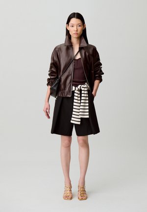 Giubbotto bomber in pelle marrone, maglia scura, gonna nera, cintura a righe e sandali con cinturini color cuoio. Design minimalista con texture lisce.