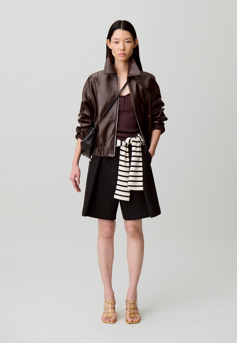 Chaqueta bomber de cuero marrón, camiseta oscura, falda negra, cinturón de rayas y sandalias de tiras en color beige. Diseño minimalista con texturas suaves.