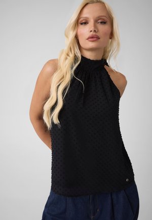 Mujer rubia con un top halter negro texturizado sin mangas y pantalones azul oscuro, de pie frente a un fondo claro y liso.
