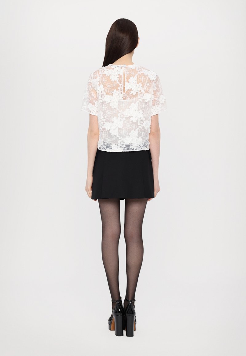 Witte kant cropped top met bloemenpatroon, gecombineerd met een zwarte flared rok. Het model draagt doorschijnende zwarte panty’s en chunky zwarte hakken.