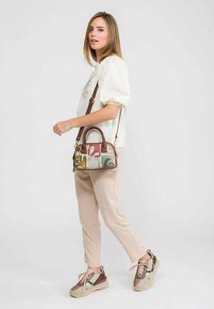 Frau trägt ein weißes Oberteil mit Puffärmeln, beige Hose, mehrfarbige Turnschuhe und eine Patchwork-Umhängetasche mit braunen Griffen und Riemen.