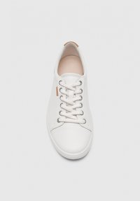 Zapatilla deportiva de cuero blanco con cordones blancos, ojales metálicos y una pequeña etiqueta marrón en el lateral, mostrada desde una vista superior sobre un fondo gris.