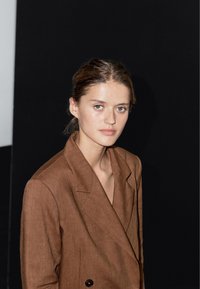 Brązowa lniana marynarka z podwójnym zapięciem, klapami z wycięciem i teksturowanym wykończeniem. Modelka ma proste włosy i neutralny wyraz twarzy.