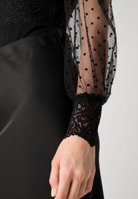 Abito nero con maniche trasparenti a pois e polsini in pizzo. Il tessuto è liscio con una finitura opaca, e il pizzo presenta un motivo intrecciato intricate.