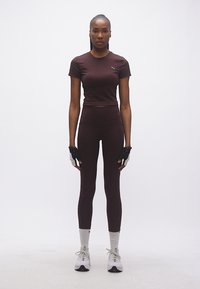 Γυναίκα όρθια, φορώντας συνδυασμένο σκούρο μπορντό αθλητικό σύνολο, γάντια χωρίς δάχτυλα, λευκές κάλτσες και λευκά αθλητικά παπούτσια, μπροστά σε απλό λευκό φόντο.