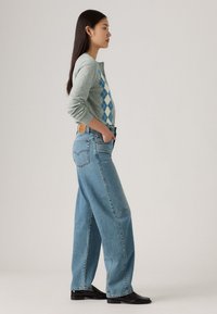 Hall nagu cardigan sinise rombimustriga, koos lohvaka sinise teksapükste paariga. Mudel seisab külje pealt, käed taskutes ja pikad tumedad juuksed.