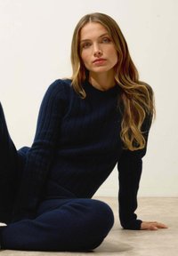 Pull en maille côtelée bleu marine à manches longues et col haut, associé à un pantalon en maille assorti, porté par une silhouette assise sur un fond clair.