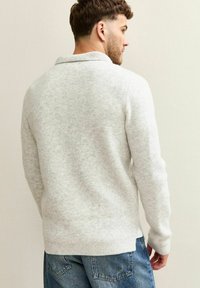 Maglione grigio lavorato a maglia con colletto ripiegato, polsini e orlo a coste. La texture sembra morbida e accogliente, indossato con jeans in denim blu.