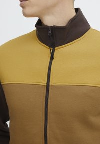 Farbblockierte Zip-Jacke aus braunem und senfgelbem Fleece-Material mit hohem Kragen und schwarzem Reißverschlussdetail.