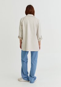 Personne aux longs cheveux bruns portant une chemise beige à manches longues et un jean bleu ample, se tenant face à un fond blanc uni.