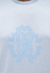 Ανοιχτό μπλε μπλουζάκι με ένα διακριτικό, περίτεχνο μοτίβο μονόγραμμα που εναλλάσσεται, τοποθετημένο στο κέντρο του στήθους.