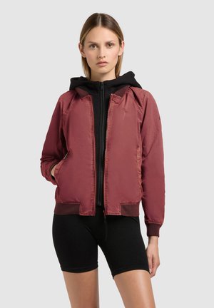 Žena v čiernej mikine s kapucňou a vínovočervenej bomber bunde s rukami v vreckách, stojaca pred jednoduchým sivým pozadím.