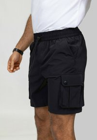 Shorts cargo noirs avec une taille élastique, détail de cordon de serrage, deux poches latérales et une poche cargo à bouton sur la cuisse.