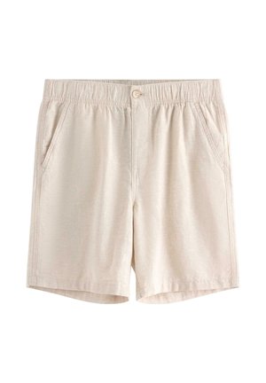 Teenagedreng stående med hænderne i lommerne iført en blå kortærmet skjorte, hvid T-shirt, beige shorts og sorte sandaler.