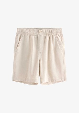 Nastolatek stoi z rękami w kieszeniach, ubrany w niebieską koszulę z krótkim rękawem, białą koszulkę, beżowe szorty i czarne sandały.