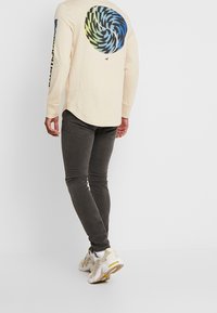 Camiseta de manga larga beige con un gráfico de espiral colorido en la parte posterior y texto negro en las mangas, combinada con jeans oscuros de corte slim y zapatillas.