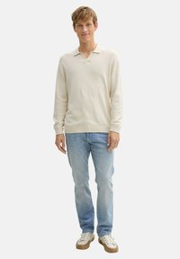 Beige hosszú ujjú pulóver, galléros nyakkal, világoskék farmernadrággal és fehér, barna talpú sportcipőkkel, egy sima háttér előtt állva.