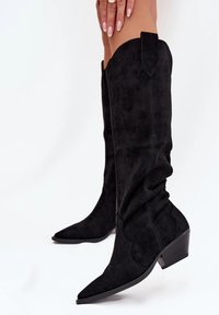 Bottes noires en daim jusqu'aux genoux avec un bout pointu, des détails brodés et un talon modeste. Présente un design flou avec des coutures accentuées.