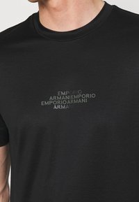 Svart t-shirt med kort ärm och rund halsringning, prydd med en upprepande logotyp i olika nyanser av grått. Slätt bomullsmaterial.