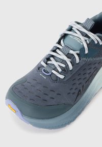 Blå atletisk sko med hvite og grå snorer, ventilert overdel, tykk såle og Vibram-logo på tåhetten.