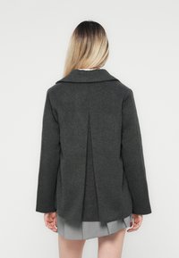 Manteau en laine gris foncé avec un col cranté, des manches longues et un détail de pli au dos, associé à une jupe plissée gris clair.