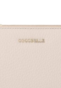 Světle růžová kožená peněženka s texturou zrnité kůže. Obsahuje zlatou embosovanou značku s nápisem "Coccinelle." Jednoduchý design, bez dalších akcentů.