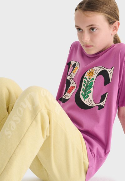 Ung person, der sidder, iført lilla T-shirt med blomstermotiv "BC" og lysegule bukser med svag tekst på benet.