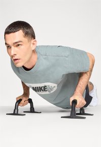 Nike t-shirt i lys blå med trykt logo, parret med sorte push-up stænger. Model udfører en push-up på en ren hvid baggrund.