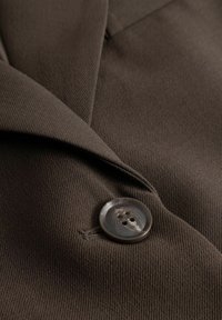 Blazer de tela marrón con un cuello con muesca y un botón circular oscuro de cuatro agujeros, mostrando una superficie texturizada.