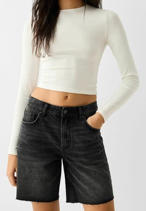 White long-sleeve cropped top paired with black denim shorts, featuring a frayed hem and five pockets, worn with hands in pockets.