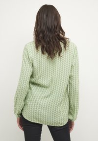 Mujer con cabello castaño ondulado, vestida con una blusa de manga larga de un patrón verde claro y pantalones negros, de espaldas contra un fondo sencillo.