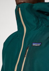 Tükist vett tõrjuv teal värvi jope kõrge kraega, beež zipp ja Patagonia logo. Omab siledat tekstuuri ja reguleeritavat kapuutsit.