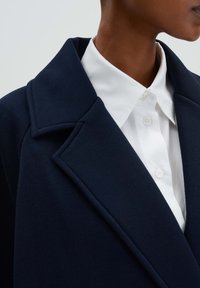 Marineblauer Wollmantel mit breitem Revers, kombiniert mit einem weißen Hemd mit Kragen und Knopfverschluss, das die glatte Stoffstruktur hervorhebt.