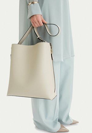 Osoba v světle modrém oblečení drží velkou krémovou koženou tašku přes rameno se zlatým zipem a tlustým popruhem po boku.