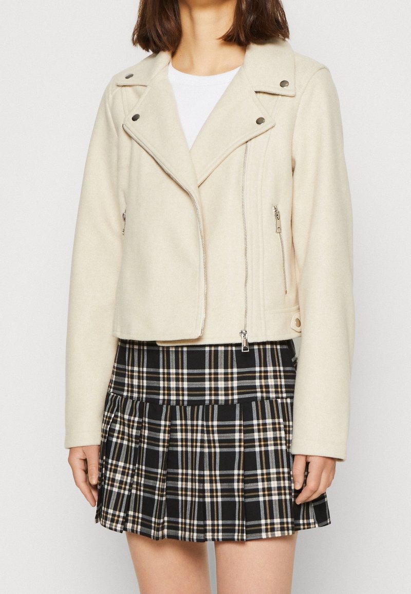 Femme portant une veste crème zippée sur une chemise blanche et une jupe plissée à carreaux noir, blanc et beige sur un fond clair.
