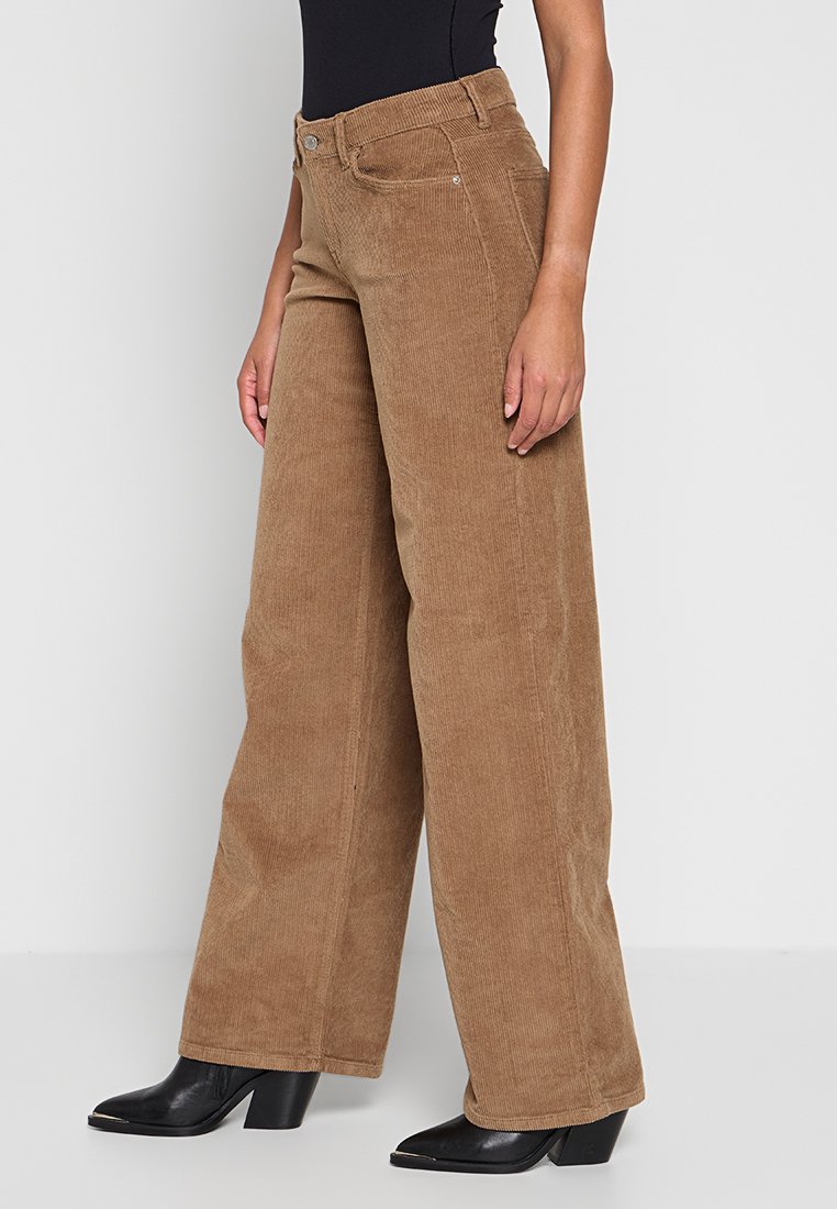 Pantaloni a gamba larga in corduroy marrone con una superficie testurizzata, vita media e tasche frontali, abbinati a stivaletti neri con tacco.