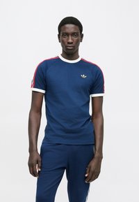 Tamsiai mėlynas medvilninis marškinėlis su balta apdaila ir raudonomis šoninėmis juostelėmis. Ant krūtinės yra mažas auksinis logotipas. Trumpomis rankovėmis, aptempto dizaino.